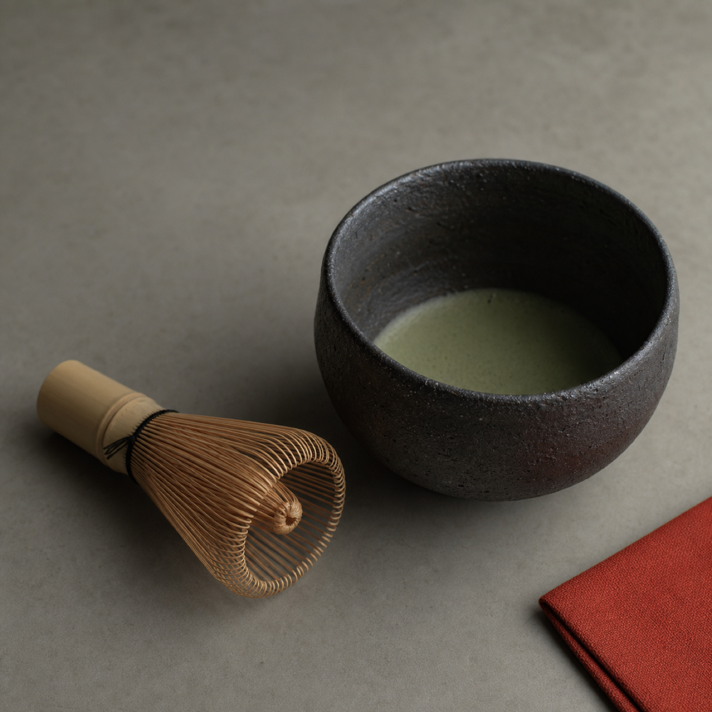 A bamboo matcha whisk beside a dark ceramic chawan of muted green matcha, soft side light, a single vermilion accent.